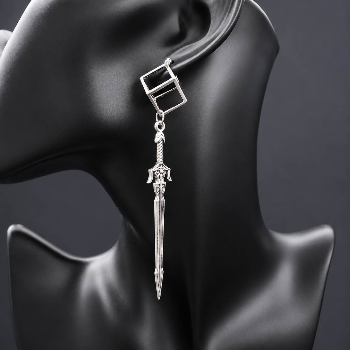 A silver dangling earring displayed on a black mannequin head against a dark gray background. The earring features a square top with a rectangular cutout, a small connecting ring, and a dangling sword-shaped charm with detailed engravings.