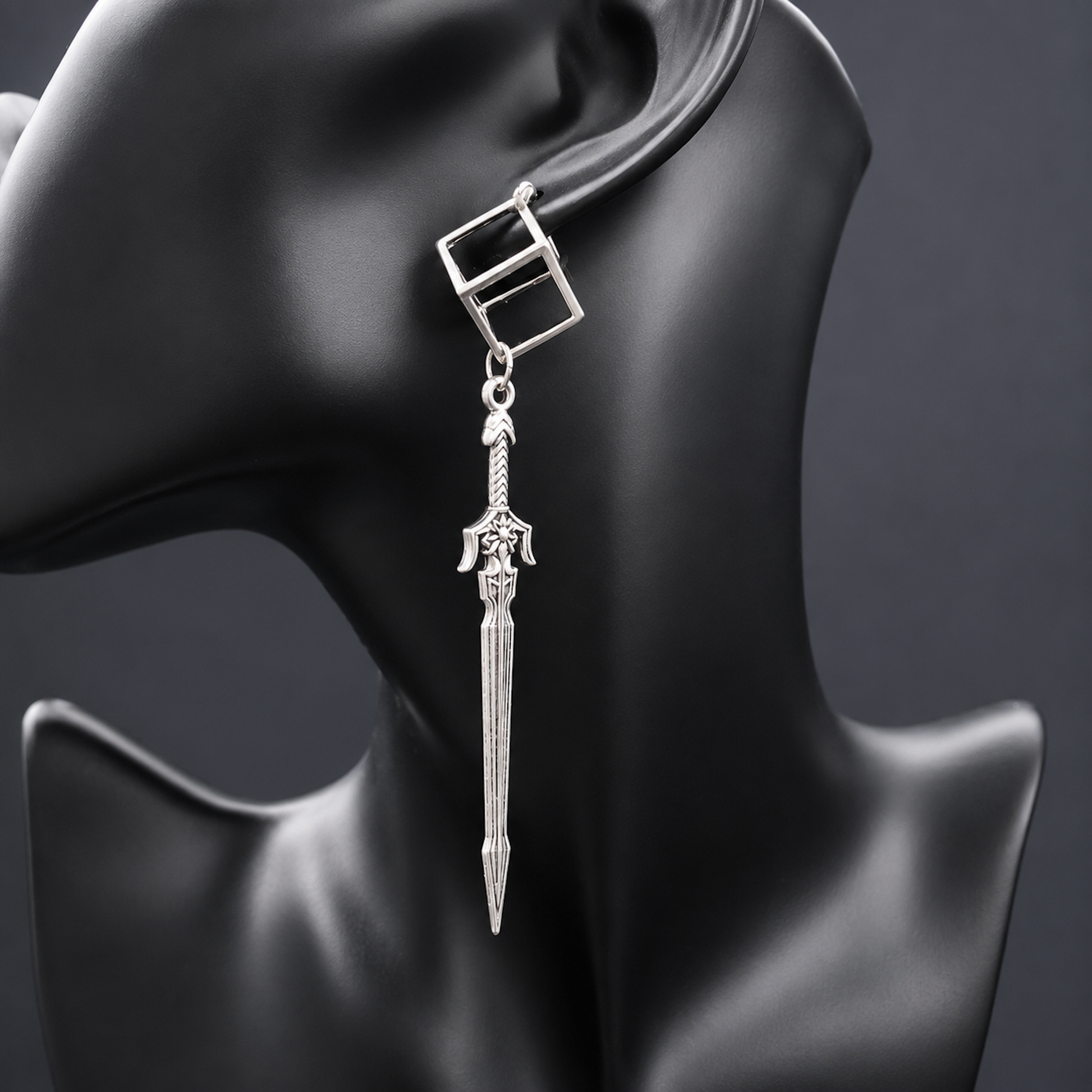 A silver dangling earring displayed on a black mannequin head against a dark gray background. The earring features a square top with a rectangular cutout, a small connecting ring, and a dangling sword-shaped charm with detailed engravings.