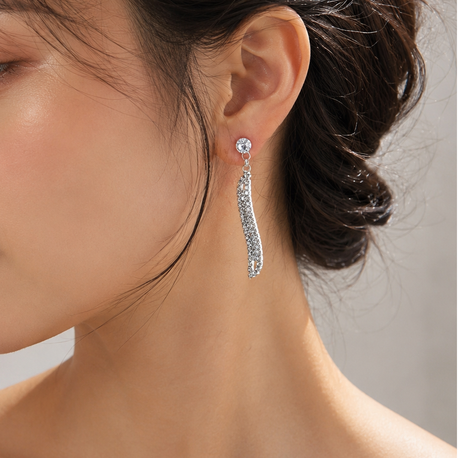 A close-up side profile of a woman wearing a long, silver-toned drop earring adorned with sparkling clear stones, set against a soft neutral background.