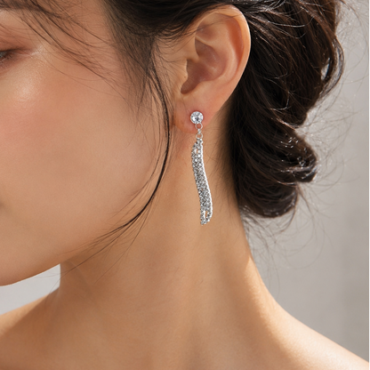 A close-up side profile of a woman wearing a long, silver-toned drop earring adorned with sparkling clear stones, set against a soft neutral background.