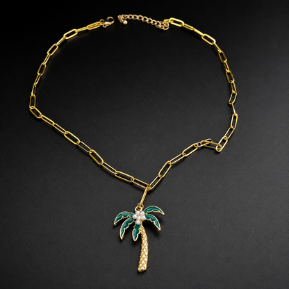 A gold paperclip chain necklace with a  palm tree pendant featuring green enamel leaves, a gold trunk, and a white faux pearl center, displayed on a dark background.