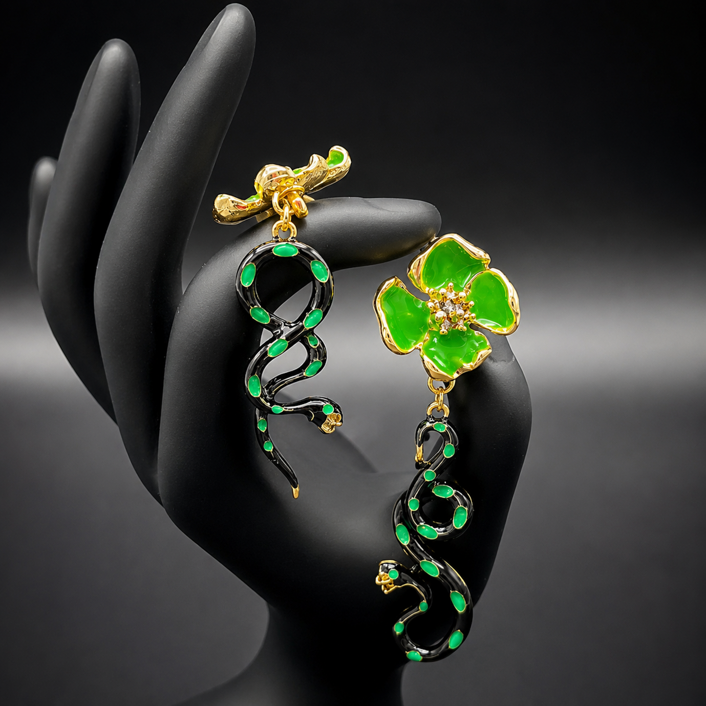 A pair of gold and green drop earrings One earring features a bright green four-petal flower with a small clear center, while the other features a black snake wrapped with green enamel and small gold details.