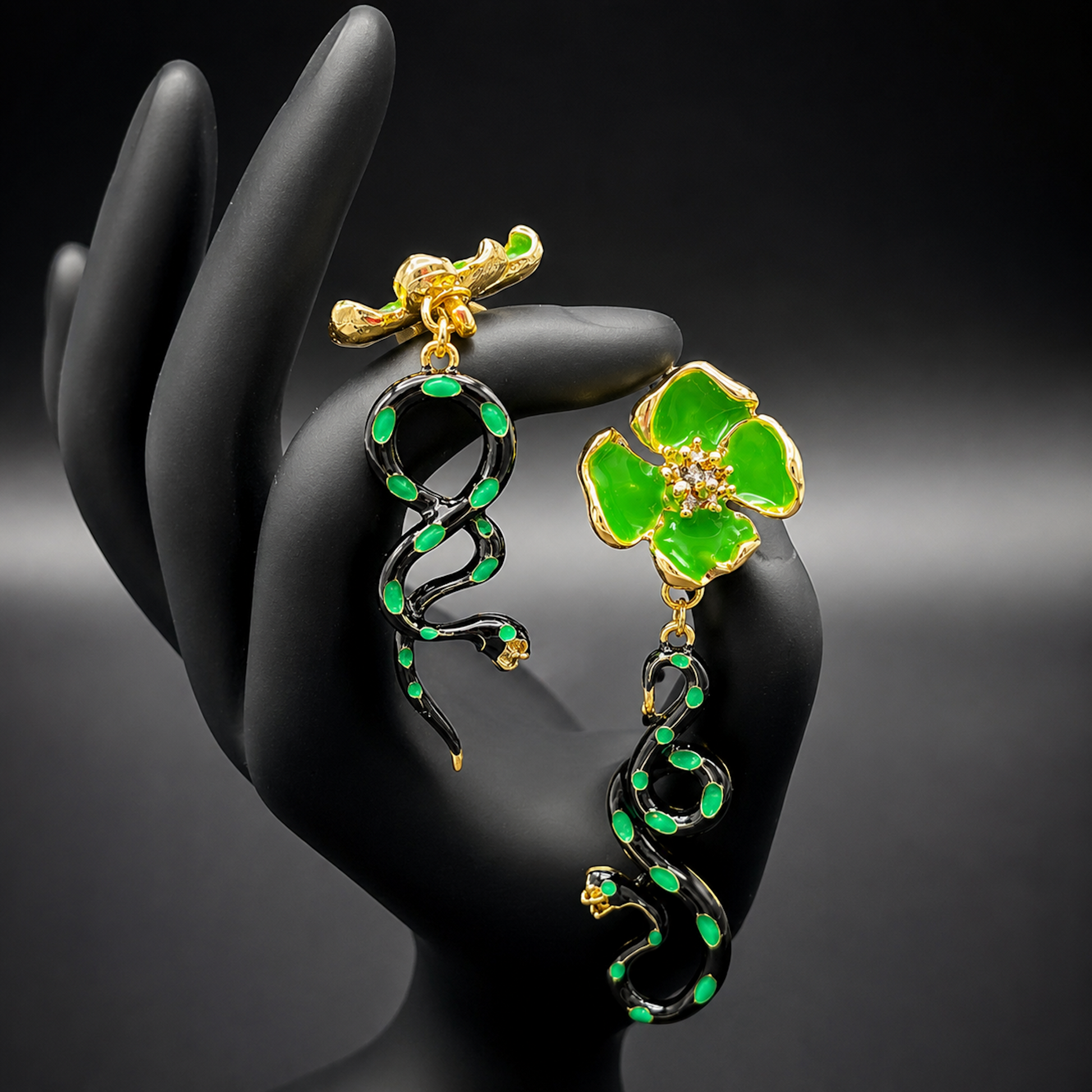 A pair of gold and green drop earrings One earring features a bright green four-petal flower with a small clear center, while the other features a black snake wrapped with green enamel and small gold details.