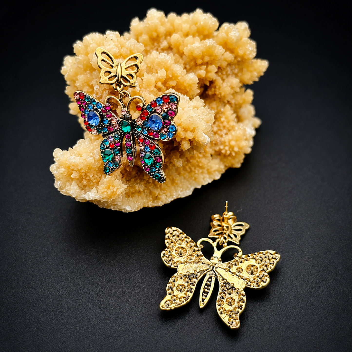 A pair of gold butterfly-shaped earrings displayed on a black background, with one earring hanging from a beige crystal cluster and the other placed below it. The butterfly wings are decorated with multicolored rhinestones.