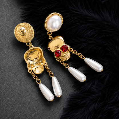 A pair of ornate gold-tone dangle earrings with white pearl-like drops, red gemstone accents, and textured gold details, displayed on a dark textured surface with black feather trim.