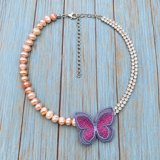 A beaded statement necklace featuring a strand of peach-pink freshwater pearls on one side, a silver-toned chain with clear rhinestones on the other, and a central butterfly applique in pink and purple. The necklace is displayed on a rustic, light blue painted wooden background.