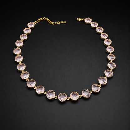 A gold-plated tennis necklace featuring round, faceted light pink gemstones, displayed on a dark textured background.