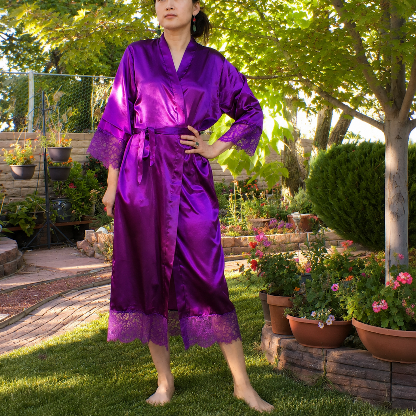 A woman stands barefoot in a sunny backyard garden wearing a shiny purple satin robe with lace trim at the cuffs and hem.