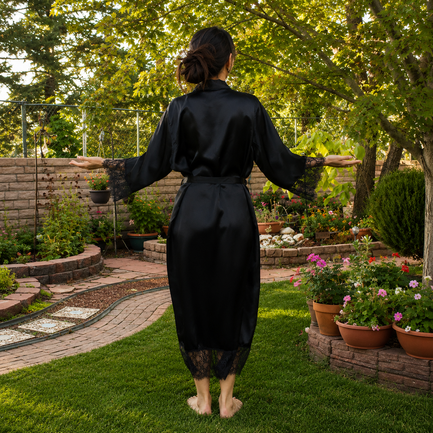 A woman stands barefoot on a grassy backyard garden path, viewed from behind, wearing a black satin-like robe with lace-trimmed sleeves and hem.