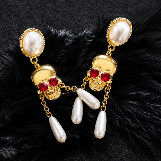 A pair of gold-toned skull dangle earrings with large white pearl oval studs at the top, red gemstone eyes, and three dangling pearl teardrop charms, displayed on a black furry surface.