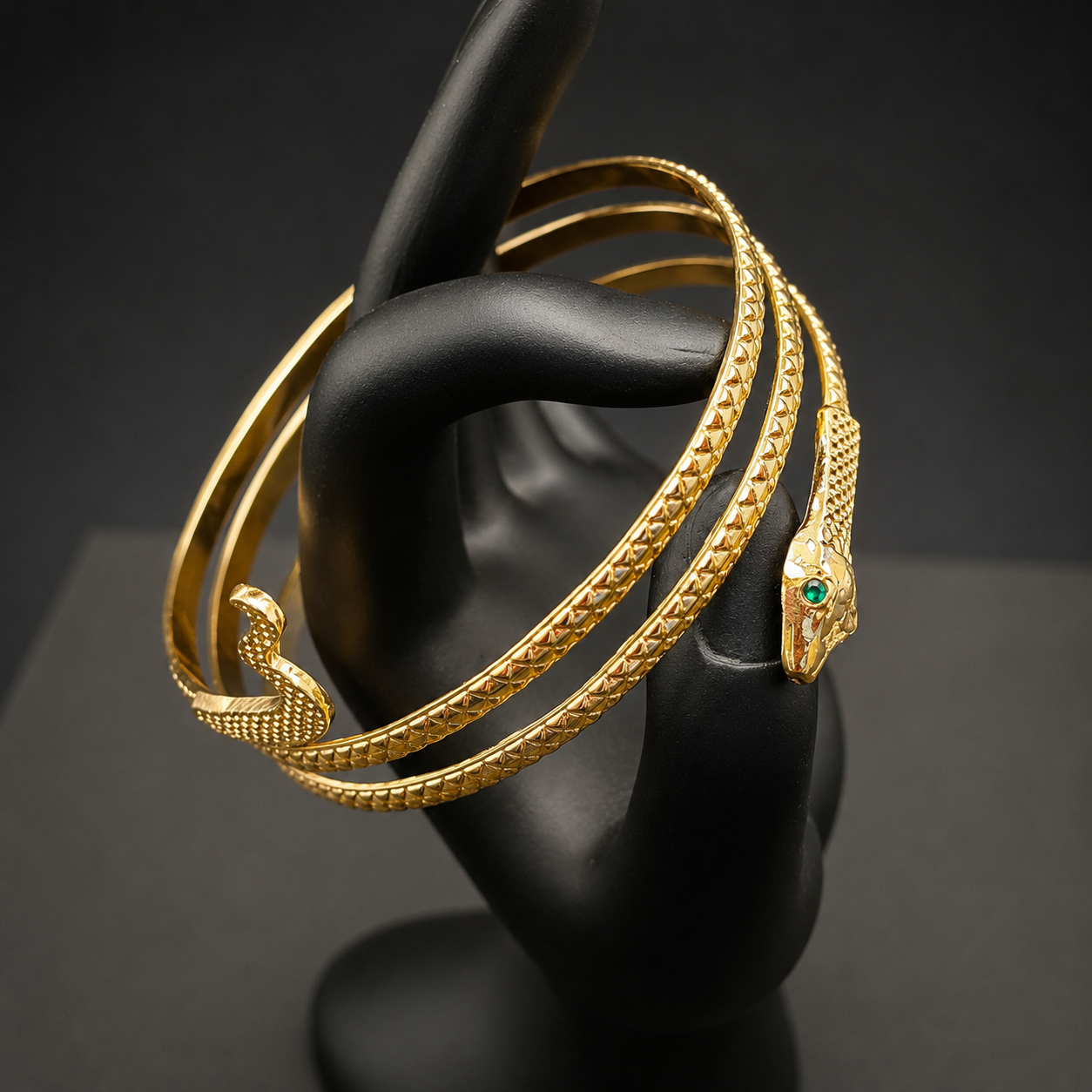 A gold snake-themed bangle bracelet displayed on a black mannequin hand. The bracelet features a textured snake body, a smaller snake head accent, and a polished snake head charm set with a small green stone.
