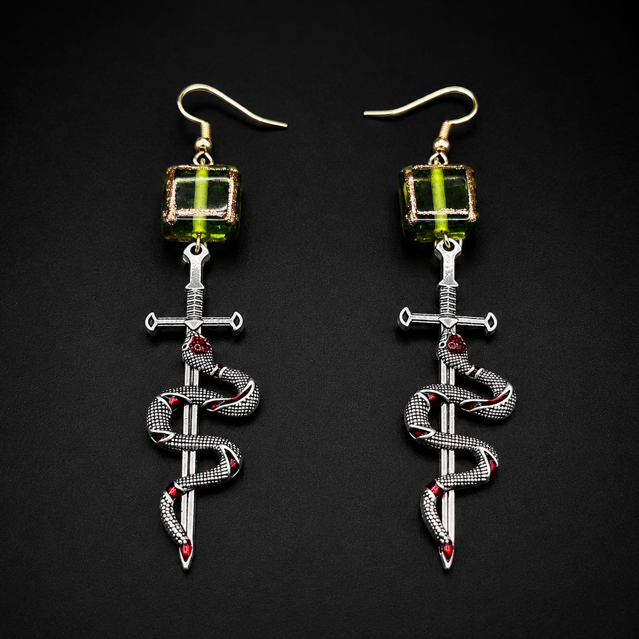 A pair of gothic-style dangle earrings on a black background. Each earring features a gold fishhook ear wire, a green faceted bead, a silver-toned dagger, and a silver snake wrapped around it with small red gemstone accents.