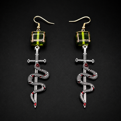 A pair of gothic-style dangle earrings on a black background. Each earring features a gold fishhook ear wire, a green faceted bead, a silver-toned dagger, and a silver snake wrapped around it with small red gemstone accents.
