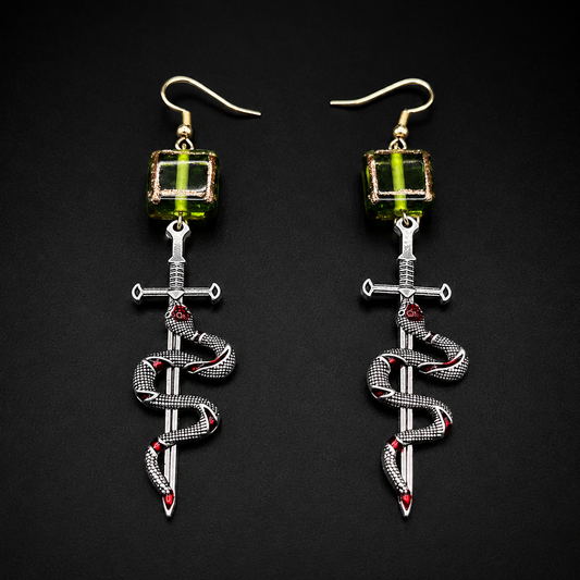 A pair of gothic-style dangle earrings on a black background. Each earring features a gold fishhook ear wire, a green faceted bead, a silver-toned dagger, and a silver snake wrapped around it with small red gemstone accents.