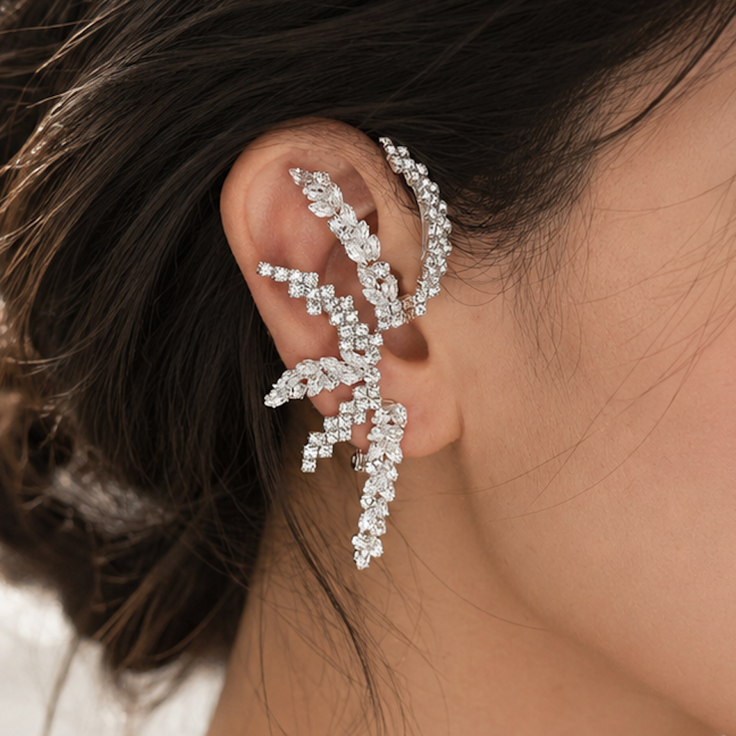A close-up of a woman wearing a delicate, silver-toned ear cuff adorned with sparkling clear rhinestones. The earring is styled on her ear, with her dark hair pulled back and a soft, bright background.