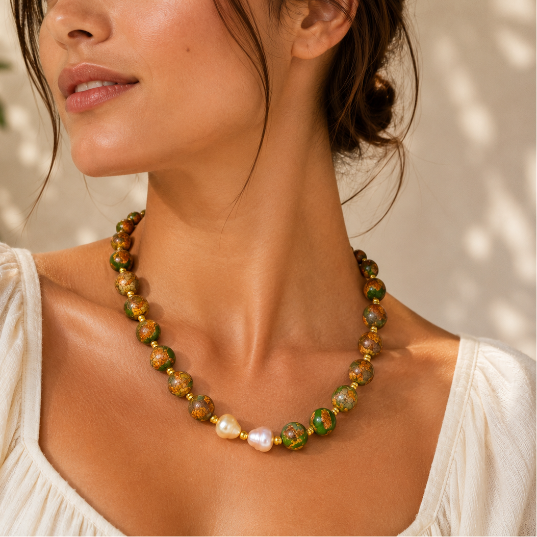 A woman wearing a beaded necklace with green, gold, and white pearls, styled with a white top against a softly lit, sun-dappled background.
