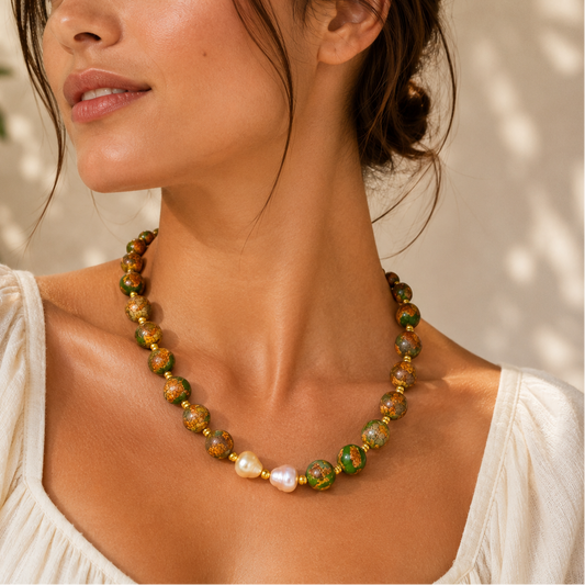 A woman wearing a beaded necklace with green, gold, and white pearls, styled with a white top against a softly lit, sun-dappled background.