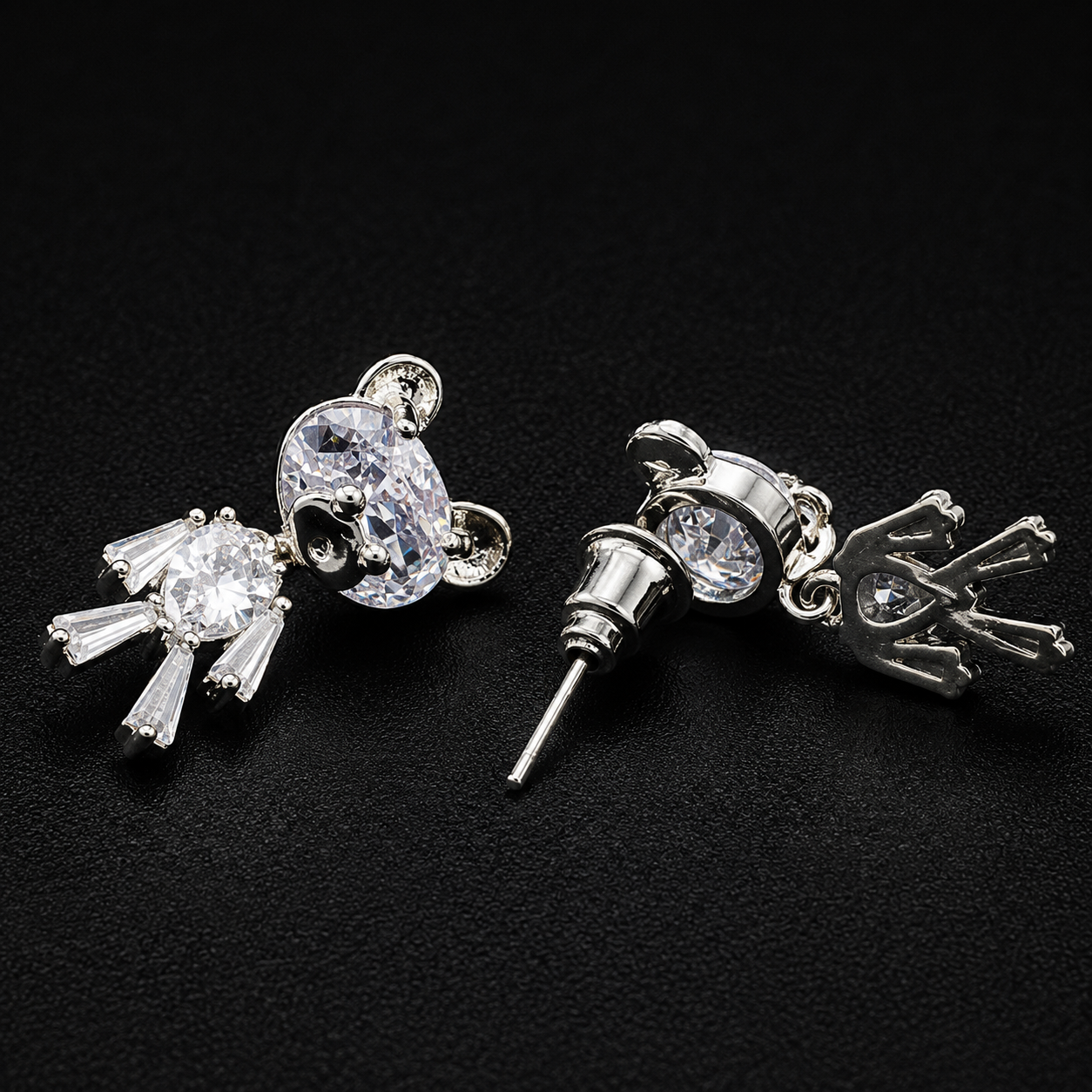 A pair of silver-toned stud earrings featuring clear crystal designs, one shaped like a mouse head and the other like a small animal with a crown, displayed on a black textured background.