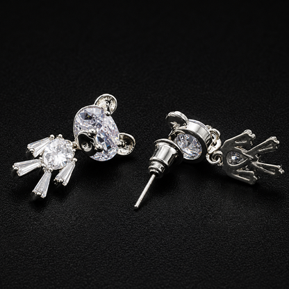 A pair of silver-toned stud earrings featuring clear crystal designs, one shaped like a mouse head and the other like a small animal with a crown, displayed on a black textured background.