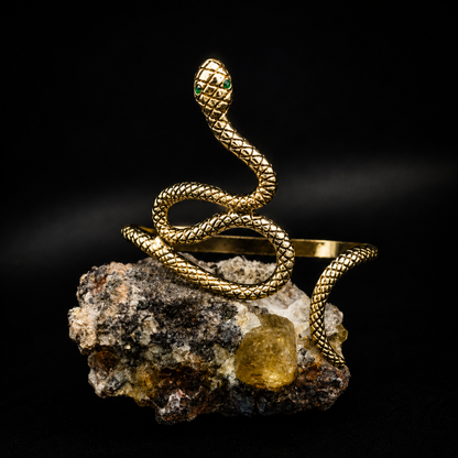 A gold snake-shaped cuff bracelet with a textured scale pattern and small green eye accents, displayed coiled on a rough mineral rock against a solid black background.